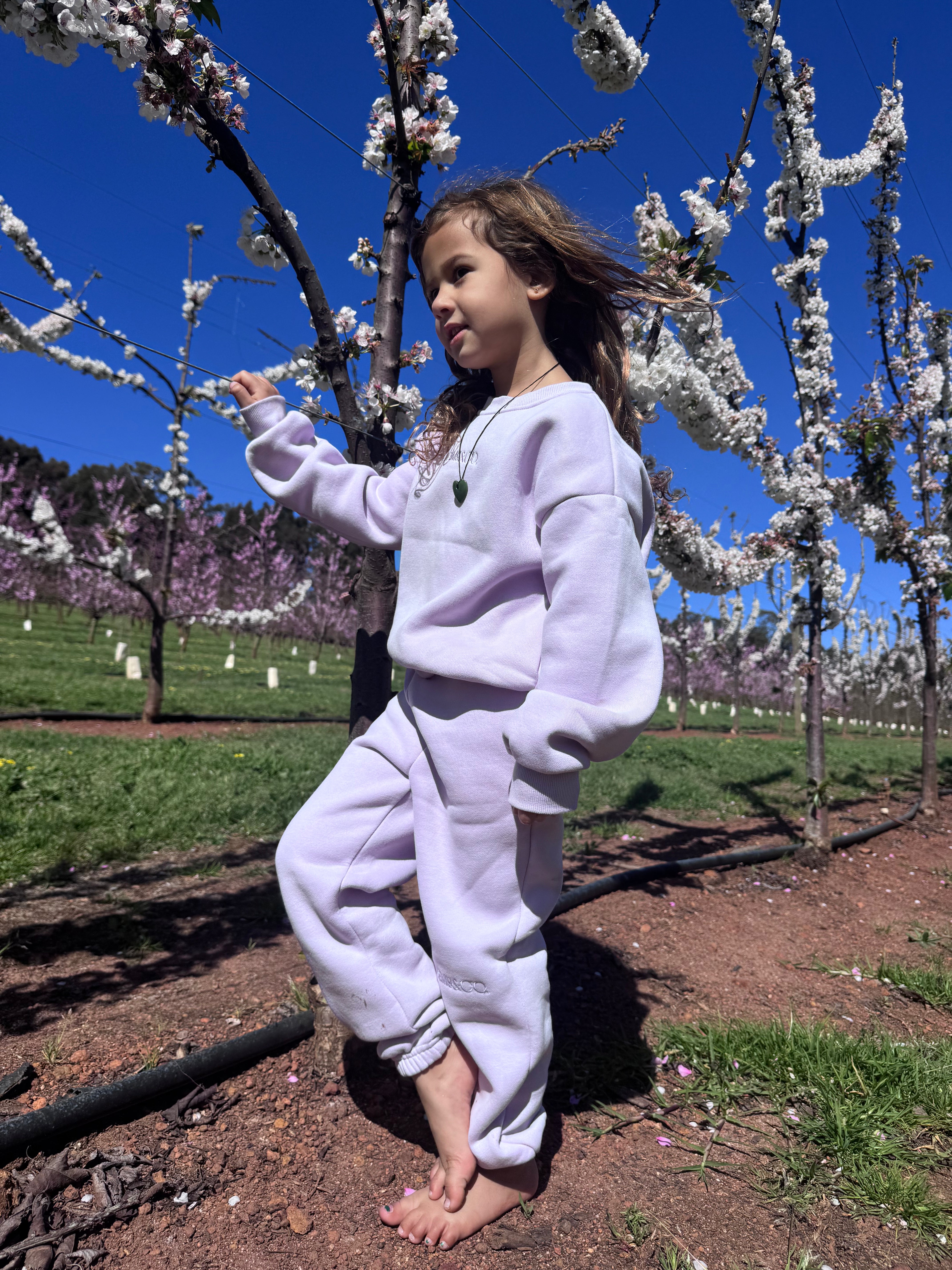 A young girl standing barefoot among blossom trees wearing the Remy & Co. Lilac crewneck sweatshirt and matching joggers.