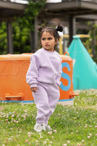 Child in a lavender outfit standing in a grassy area with playground equipment in the background.