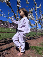 A young girl standing barefoot among blossom trees wearing the Remy & Co. Lilac crewneck sweatshirt and matching joggers.
